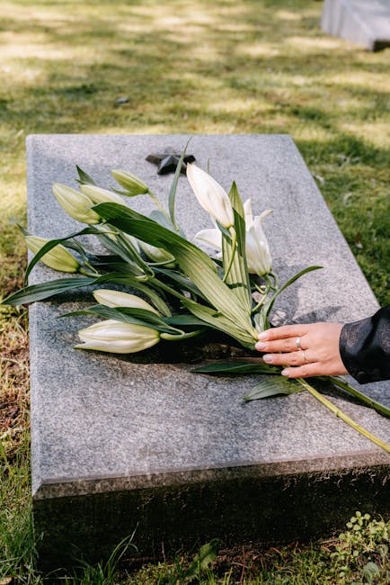 A hand with a silver ring gently places a bouquet of white lilies and lush green foliage on a polished gray stone memorial. The bouquet features multiple partially opened and closed lilies, with broad