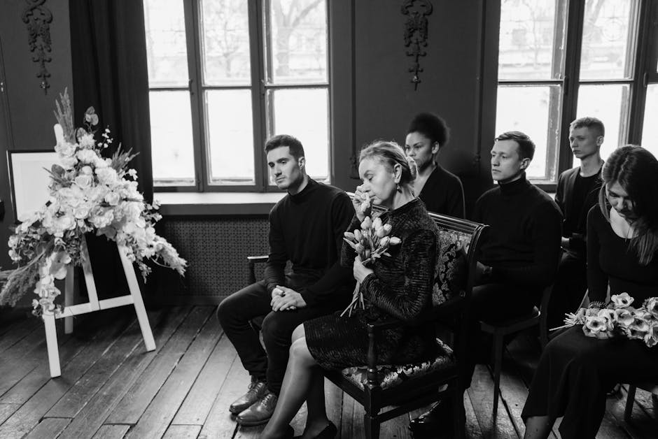 A group of six people seated indoors during a somber occasion, with a large, elegant floral arrangement on an easel to the left. The arrangement features a mix of white flowers, such as roses and lili
