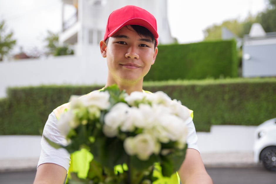 A delivery person wearing a red cap and a neon yellow vest is holding a bouquet of fresh white flowers, potentially roses or peonies, wrapped in clear and green floral paper, with some greenery visibl
