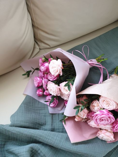 A bouquet of fresh pink and white roses with green foliage, wrapped in pale pink and lavender paper, tied with a matching pink ribbon, resting on a soft, blue-green fabric surface near a beige sofa. T